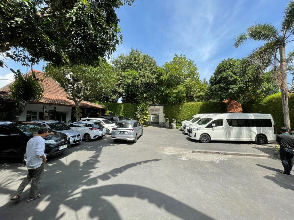 Halaman parkir yang luas dengan latar pohon hijau yang rindang. Cocok dengan konsep alam